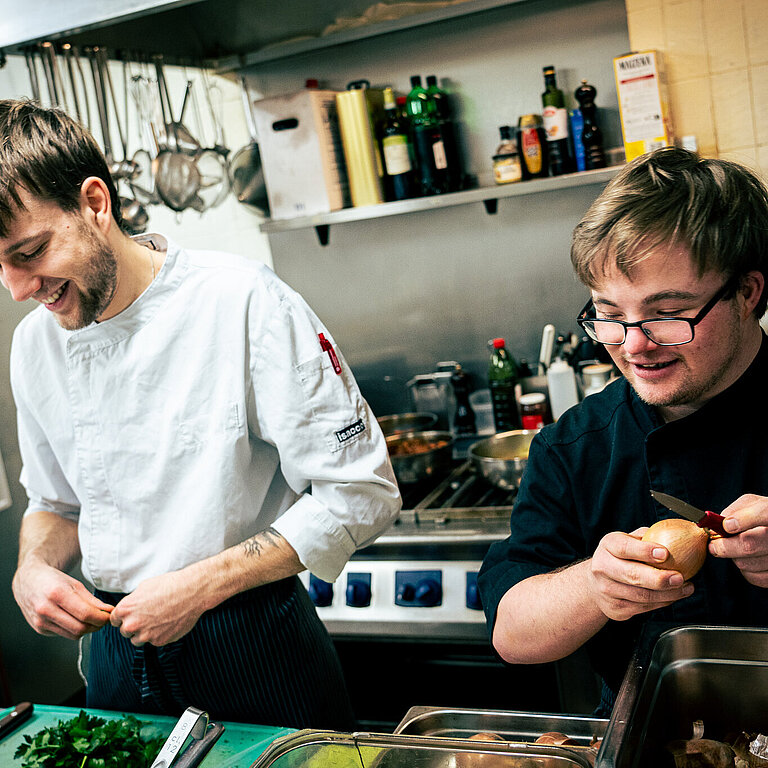 Zwei Männer in einer Restaurantküche. Ein Koch und ein Auszubildender mit Beeinträchtigung. Sie bearbeiten Gemüse, schälen Kartoffeln. 