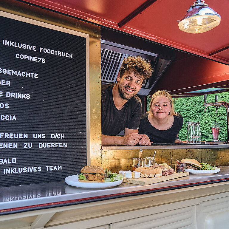 Ein Mitarbeiter und eine Mitarbeiterin mit Beeinträchtigung stehen im Foodtruck und schauen heraus. Auf der Ablage stehen Burger und Apéro.
