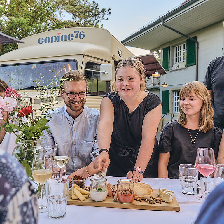 Eine Gruppe bei einem Apéro an einem Tisch. Ein Foodtruck ist im Hintergrund. 