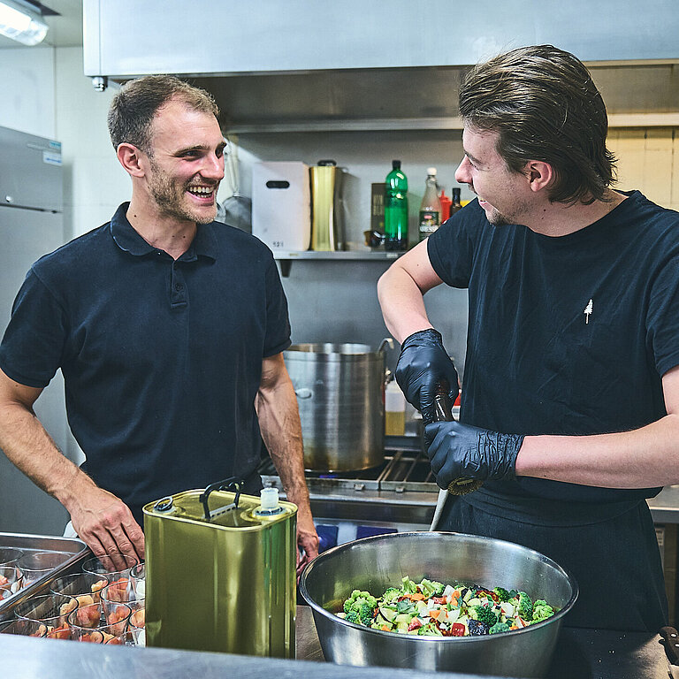 Zwei männliche Köche bei der Arbeit. Sie tragen schwarze Tshirts, lachen miteinander. In Töpfen befindet sich Gemüse. 
