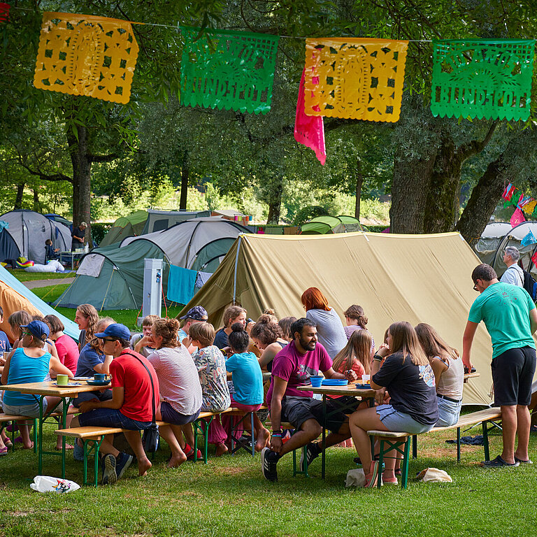Ein Zeltplatz mit vielen Zelten, vielen Kindern, die an Holztischen zusammensitzen und essen. Es hat farbige Fähnchen, die aufgehängt wurden. 