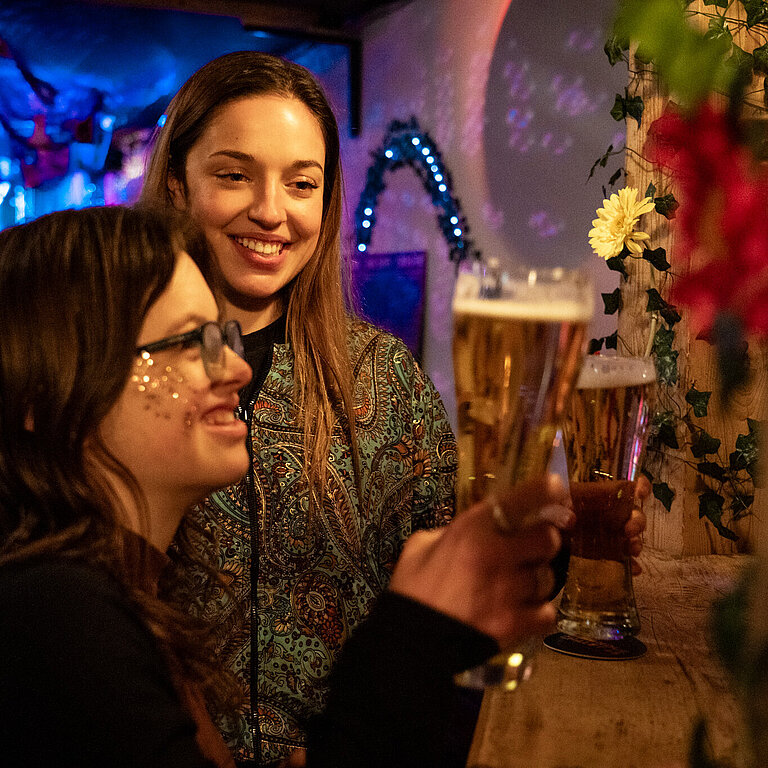 Zwei junge Frauen an einer Party an der Bar mit einem Bier in der Hand.
